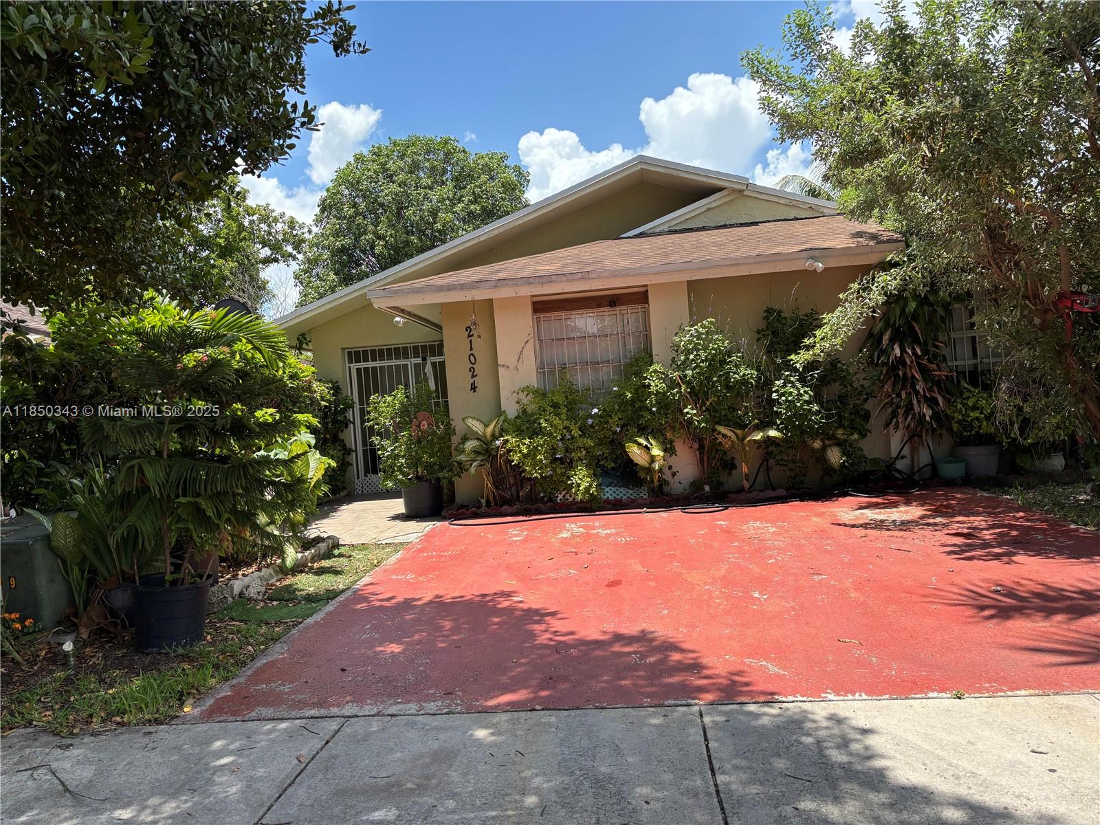 A single-story house with a red driveway and overgrown landscaping under a blue sky — 21024 SW 118th Pl, Miami FL 33177, MIAMI, FL