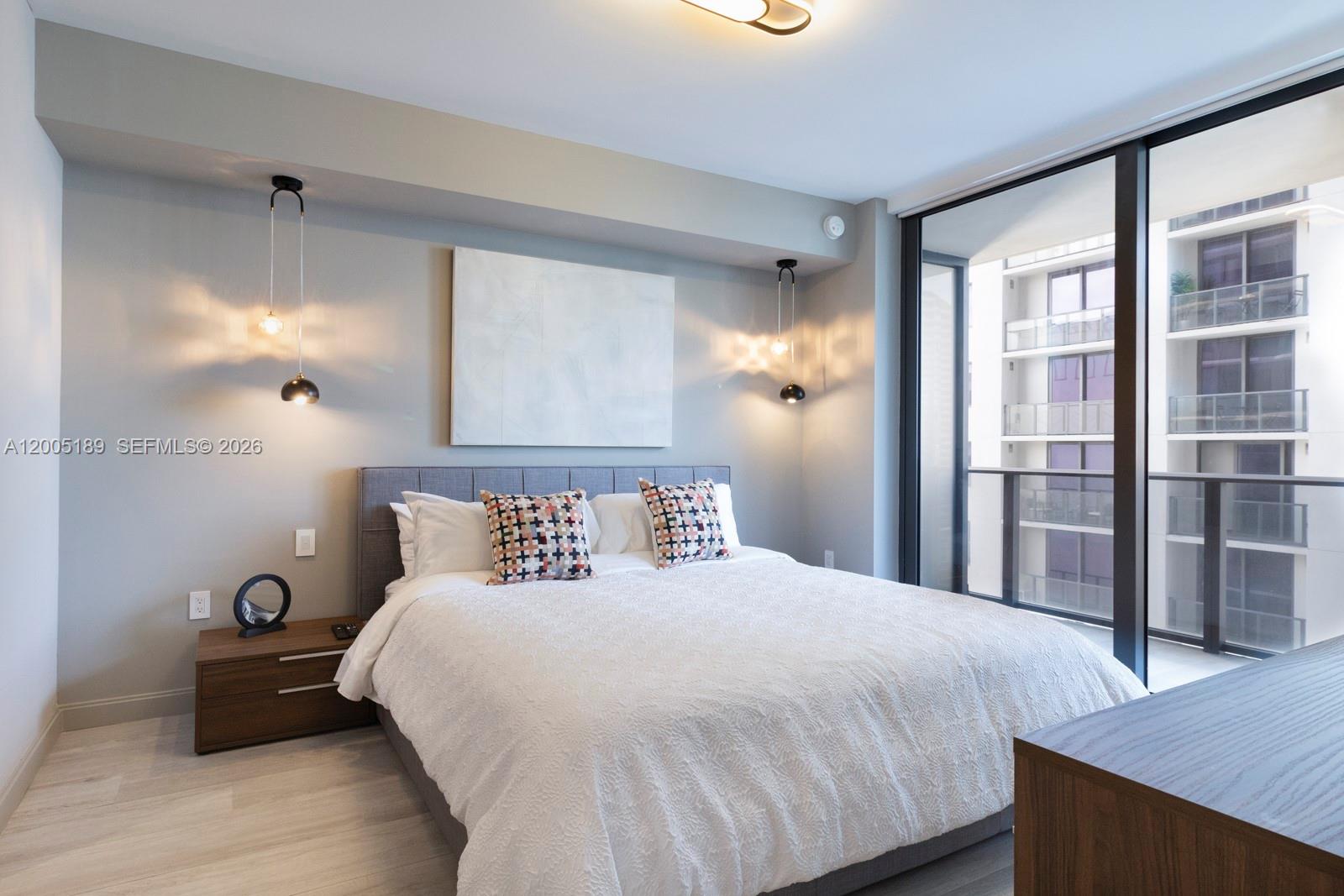 A bedroom features a gray upholstered bed, pendant lighting, and a balcony with city views — 55 SW 9th St # 3010, Miami FL 33130, MIAMI, FL