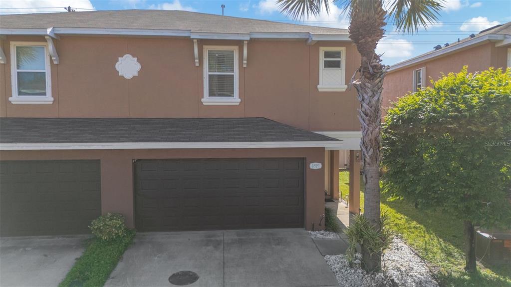 A two-story townhouse exterior features a brown stucco facade, a dark garage door, and a palm tree — 1819 SOMMARIE WAY, TARPON SPRINGS, FL