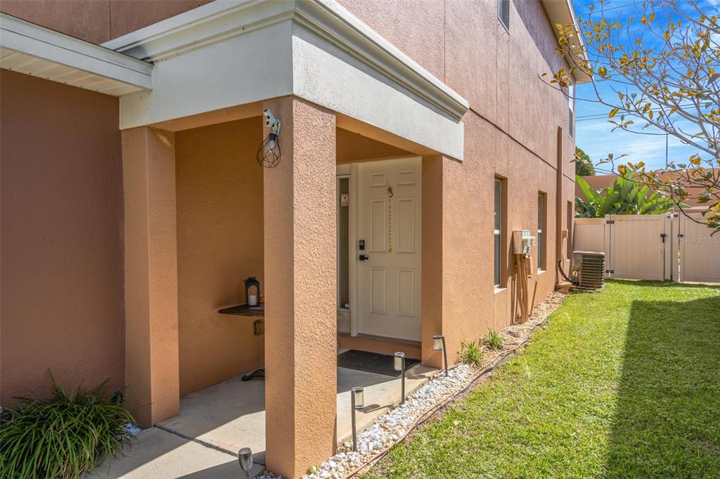 A tan stucco house exterior features a covered entryway, a white door, and a lawn — 1819 SOMMARIE WAY, TARPON SPRINGS, FL