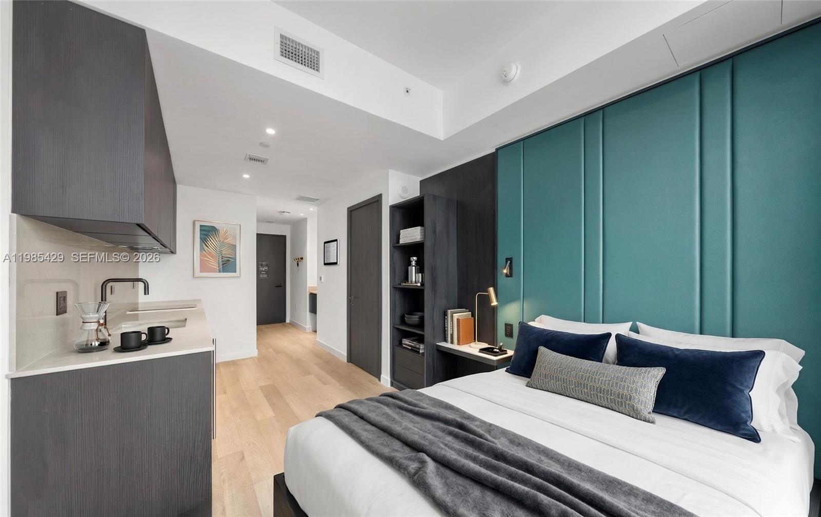 A studio apartment features a kitchenette, light wood flooring, and a teal upholstered headboard wall — 20 NE 11 ST # 2306B, Miami FL 33132, MIAMI, FL