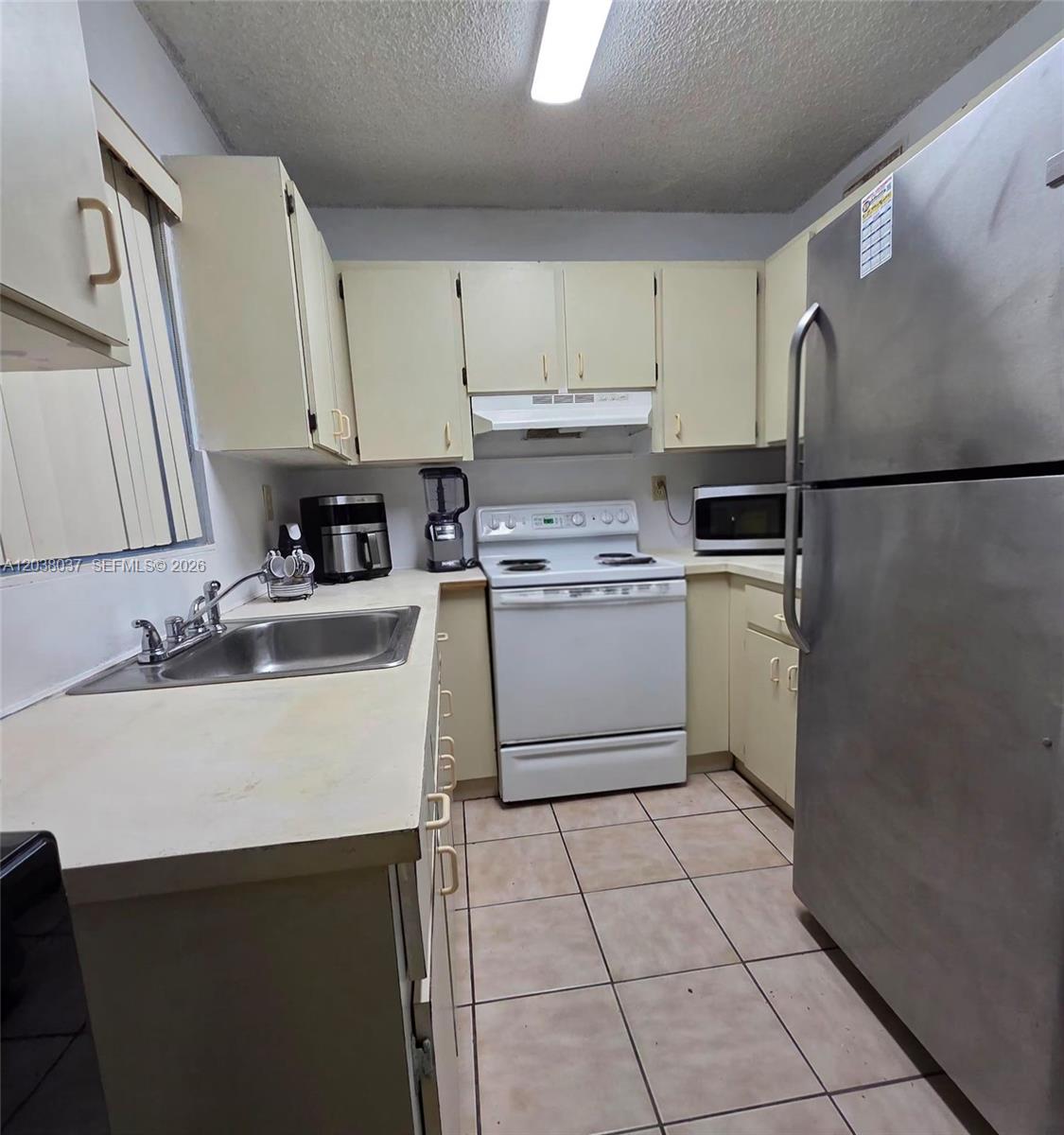 A small kitchen features light-colored cabinets, a white stove, a stainless steel refrigerator, and tile flooring — 2560 W 60th St # 104, Hialeah FL 33016, HIALEAH, FL