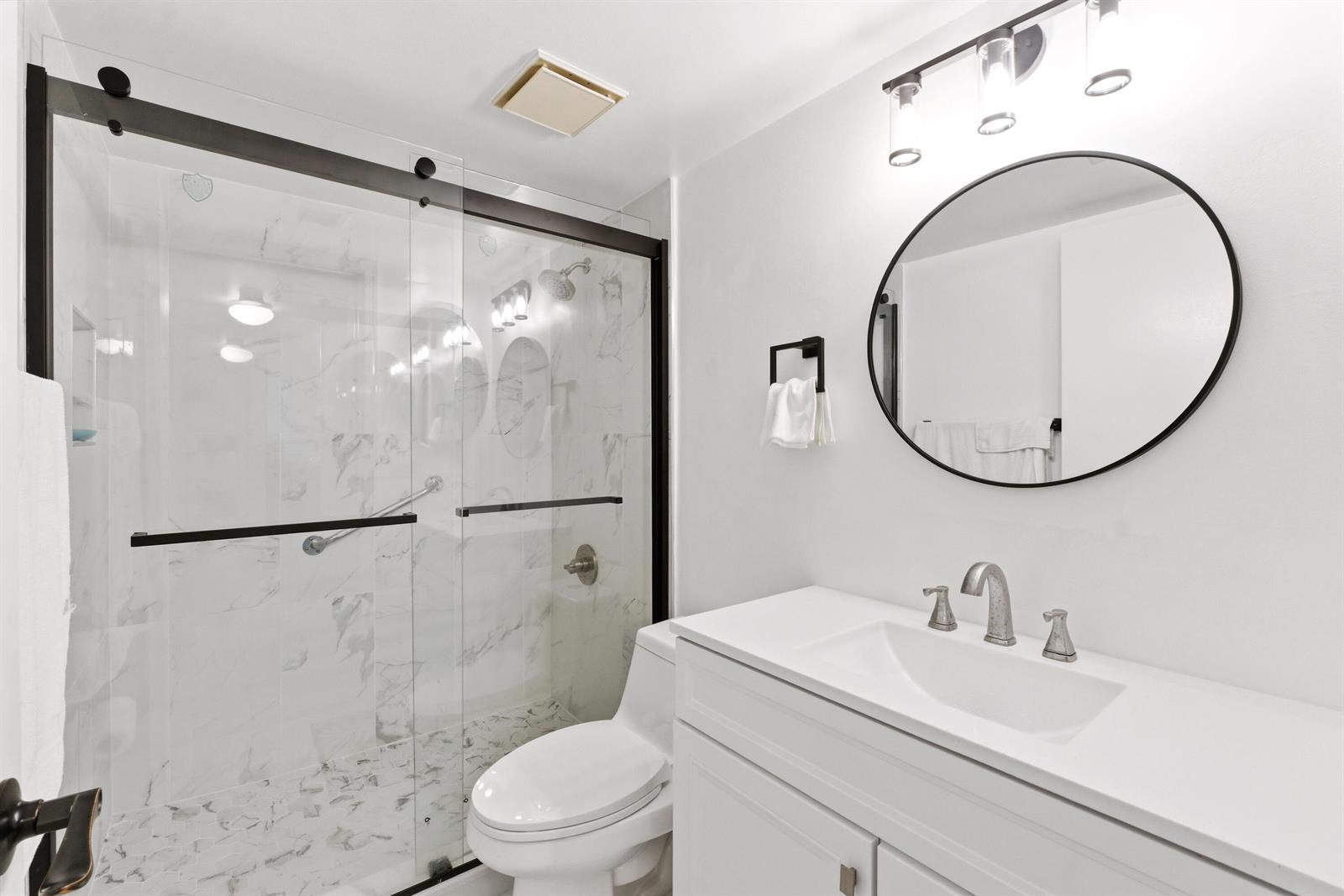 A modern bathroom features a marble-tiled walk-in shower, a white vanity, and a round mirror — 260 NW 76th Avenue # 207, Margate FL 33063, MARGATE, FL