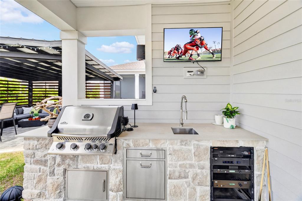 An outdoor kitchen features a stainless steel grill, sink, stone base, and wall-mounted television — 11980 SW 31ST ROAD, GAINESVILLE, FL