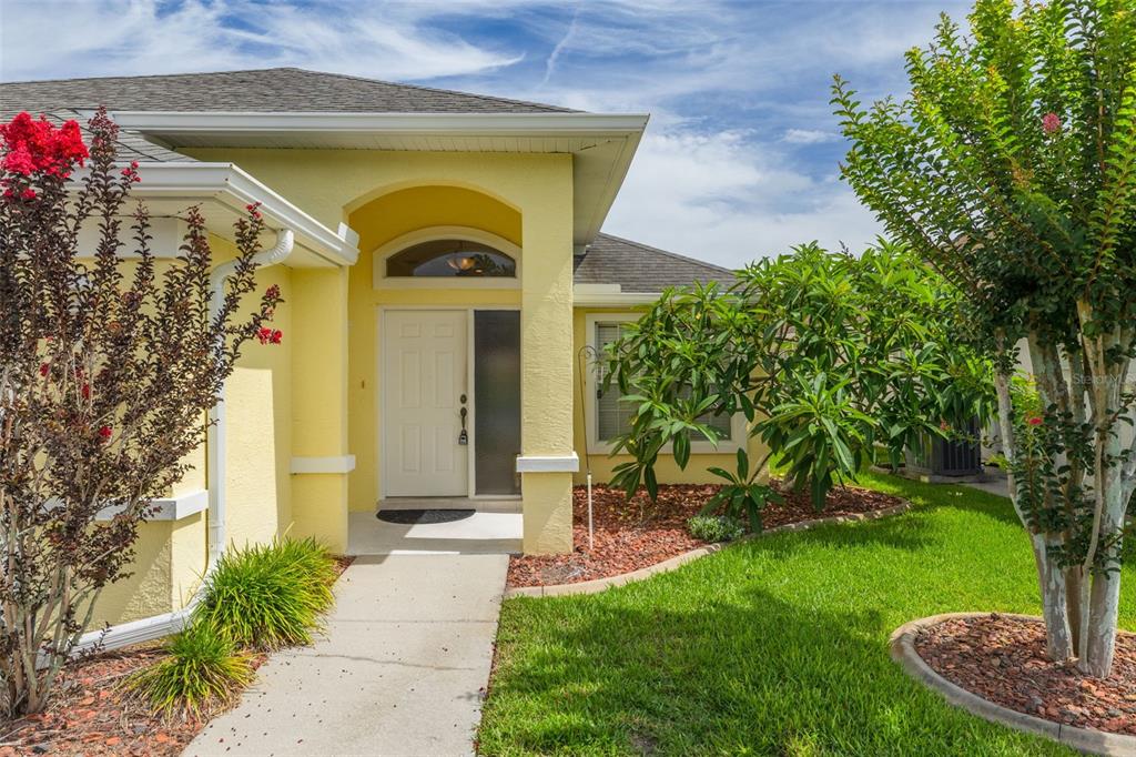 The exterior of a yellow house features a covered entryway, a white door, and landscaping — 7843 CRAIGHURST LOOP, NEW PORT RICHEY, FL