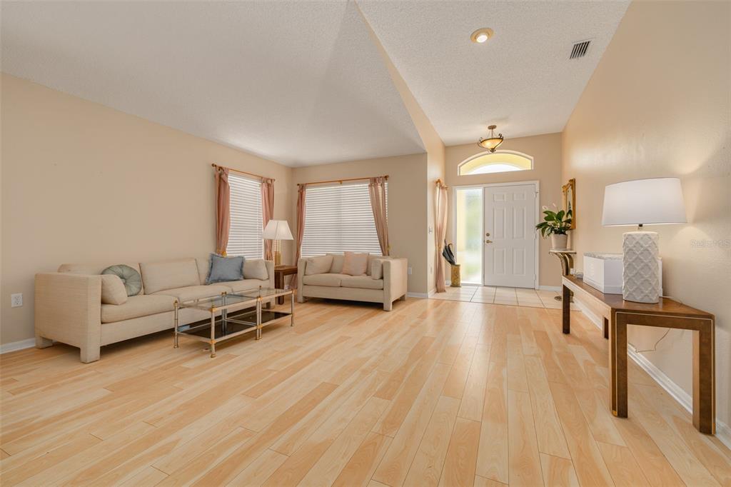 A living room with light wood flooring, beige walls, two sofas, and an entryway door — 7843 CRAIGHURST LOOP, NEW PORT RICHEY, FL