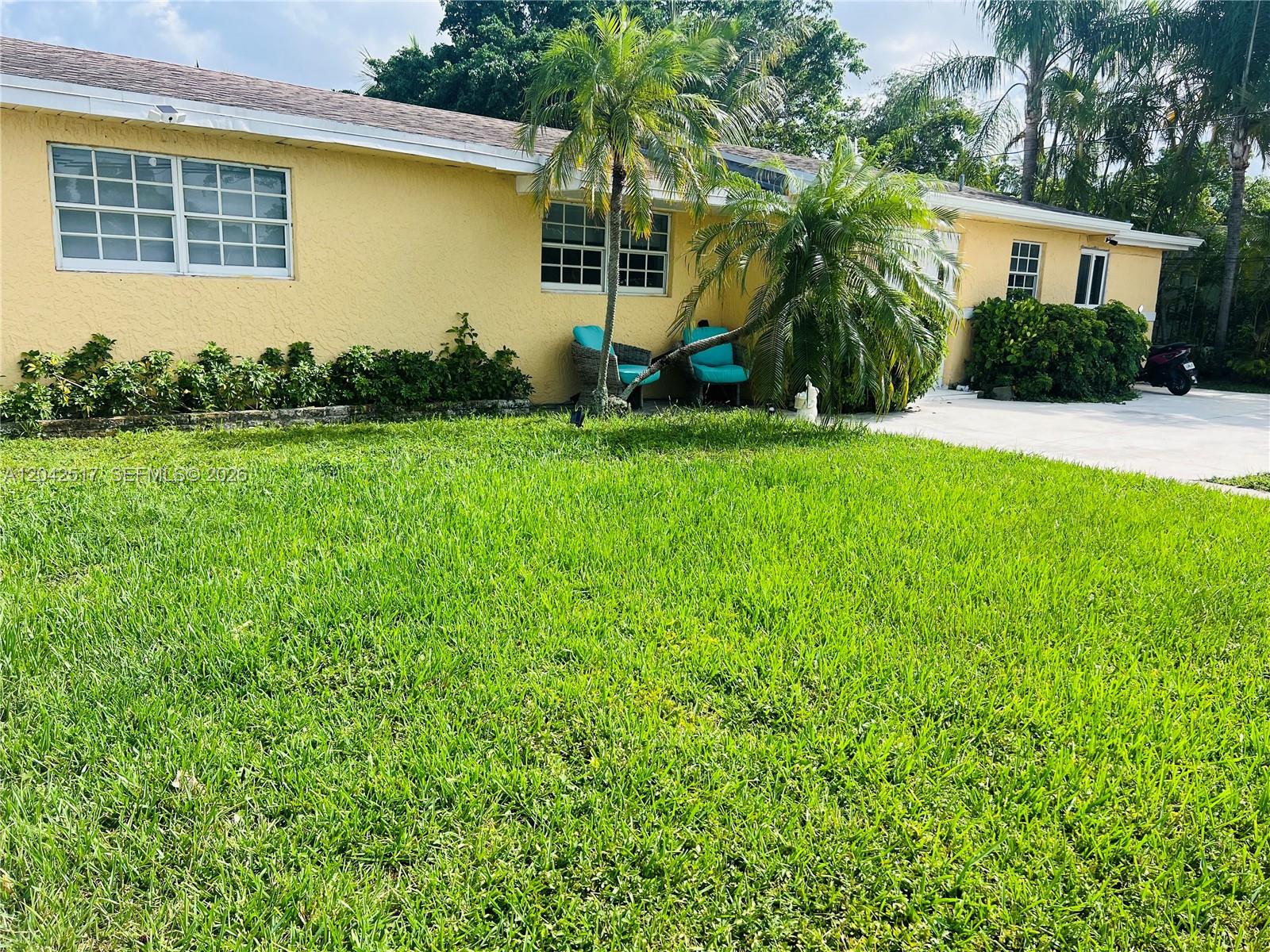 A yellow single-story house features a green lawn, palm trees, and two blue chairs — MIAMI, FL