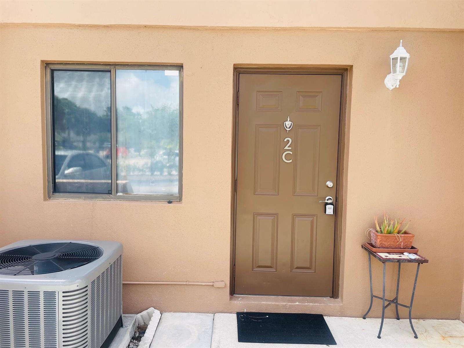 A brown exterior door labeled 2C sits next to a window, air conditioner, and plant stand — 139 Sparrow Drive # 2c, Royal Palm Beach FL 33411, ROYAL PALM BEACH, FL