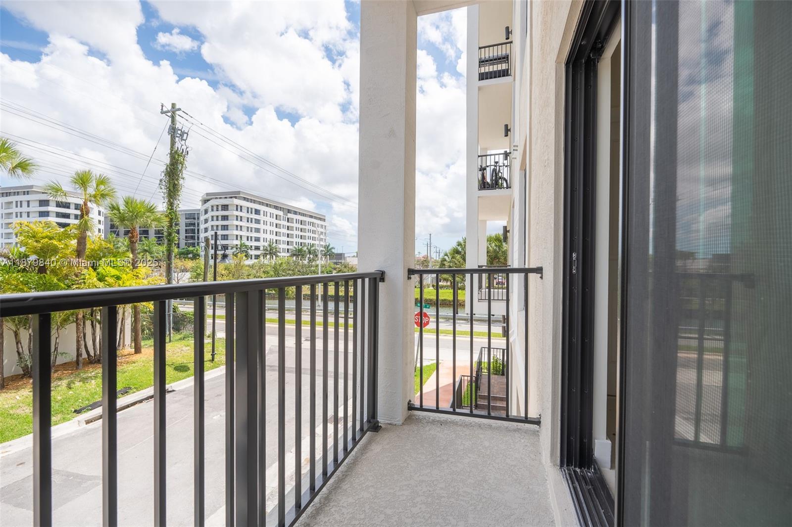 A balcony with metal railings overlooks a street, palm trees, and a nearby apartment building — 8001 NW 41st St # 208, Doral FL 33166, DORAL, FL