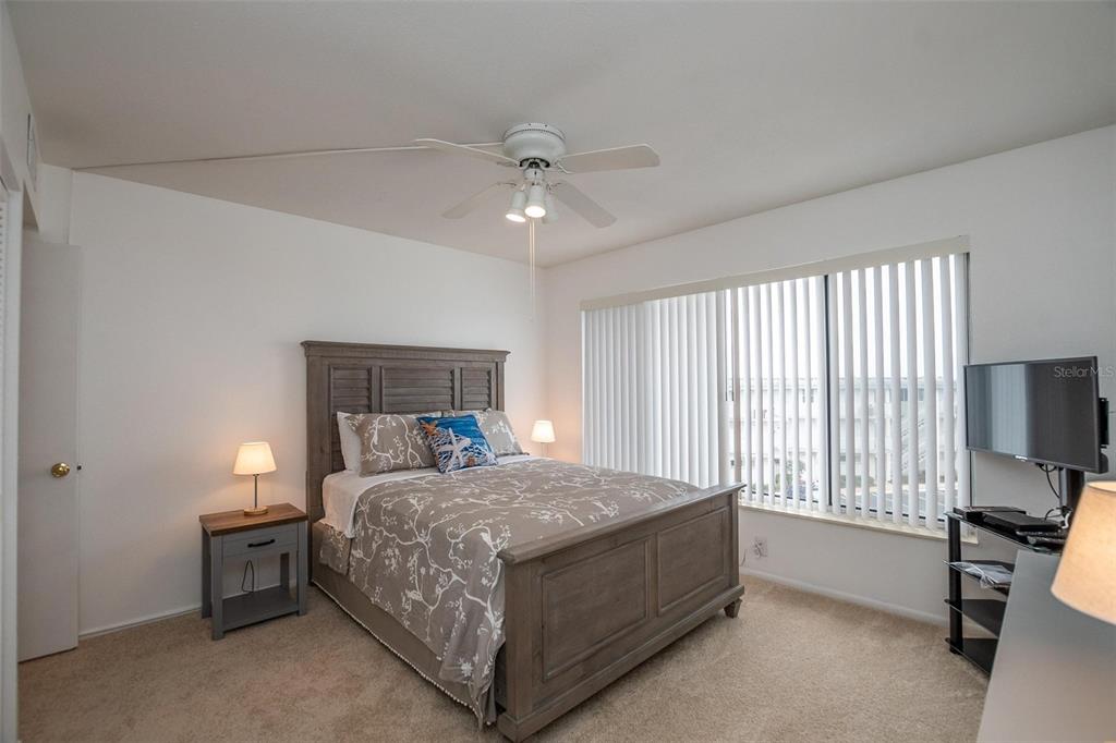 A bedroom features a wooden bed frame, a ceiling fan, and a large window with blinds — 4849 SAXON DRIVE UNIT C202, NEW SMYRNA BEACH, FL