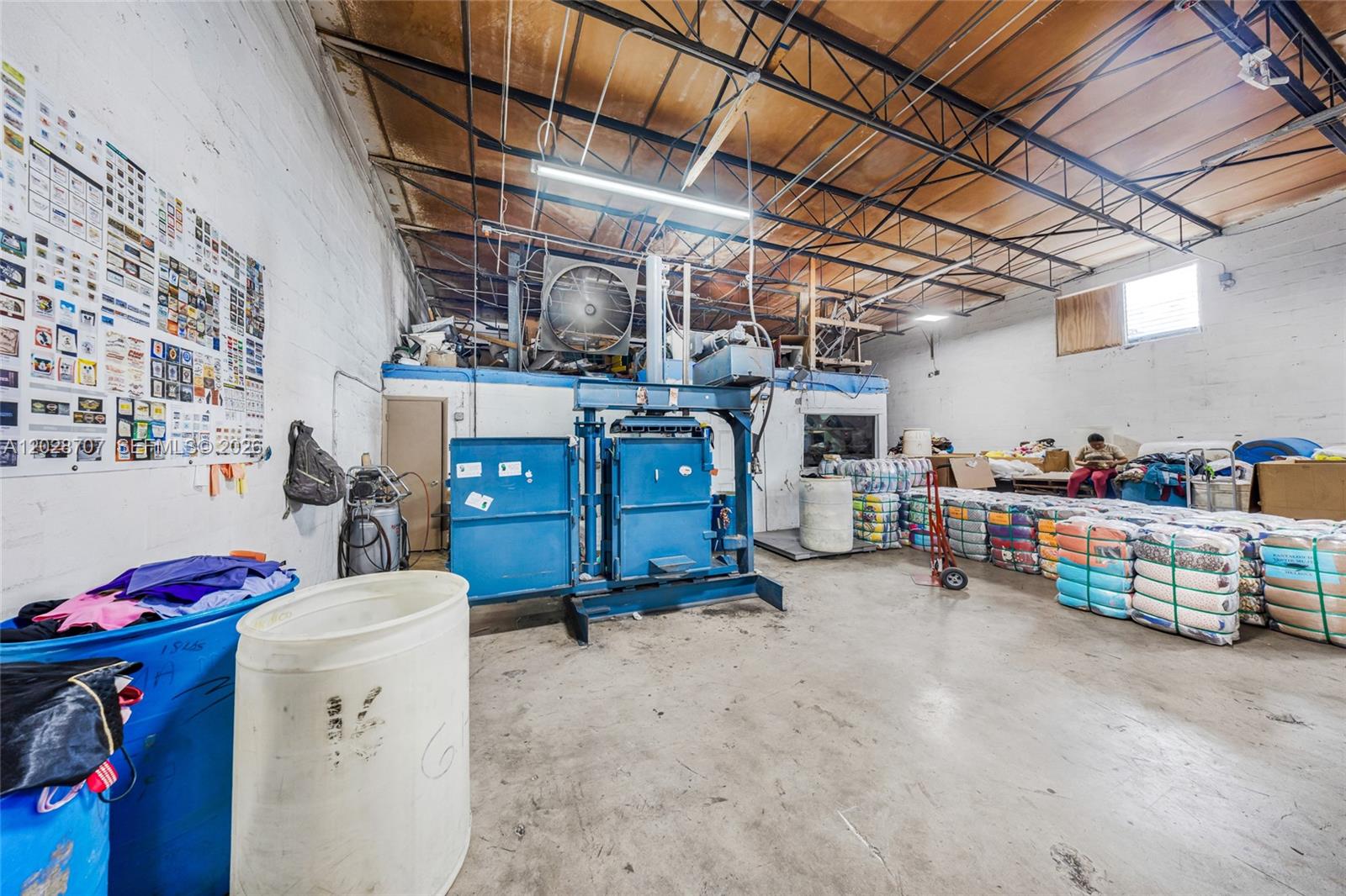 A warehouse interior features a large blue industrial baler, stacks of wrapped textiles, and concrete floors — 1085 E 27th St, Hialeah FL 33013, HIALEAH, FL