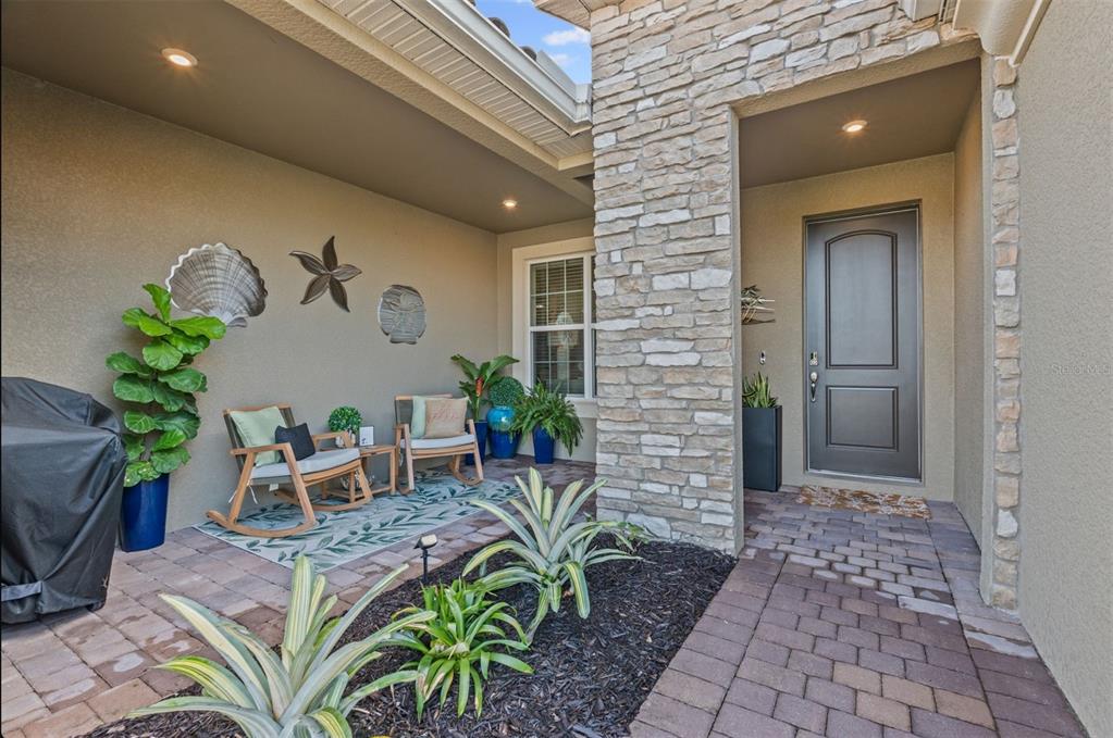 A covered front porch features a stone pillar, paver flooring, two rocking chairs, and plants — 20380 SYMPHONY PLACE, VENICE, FL