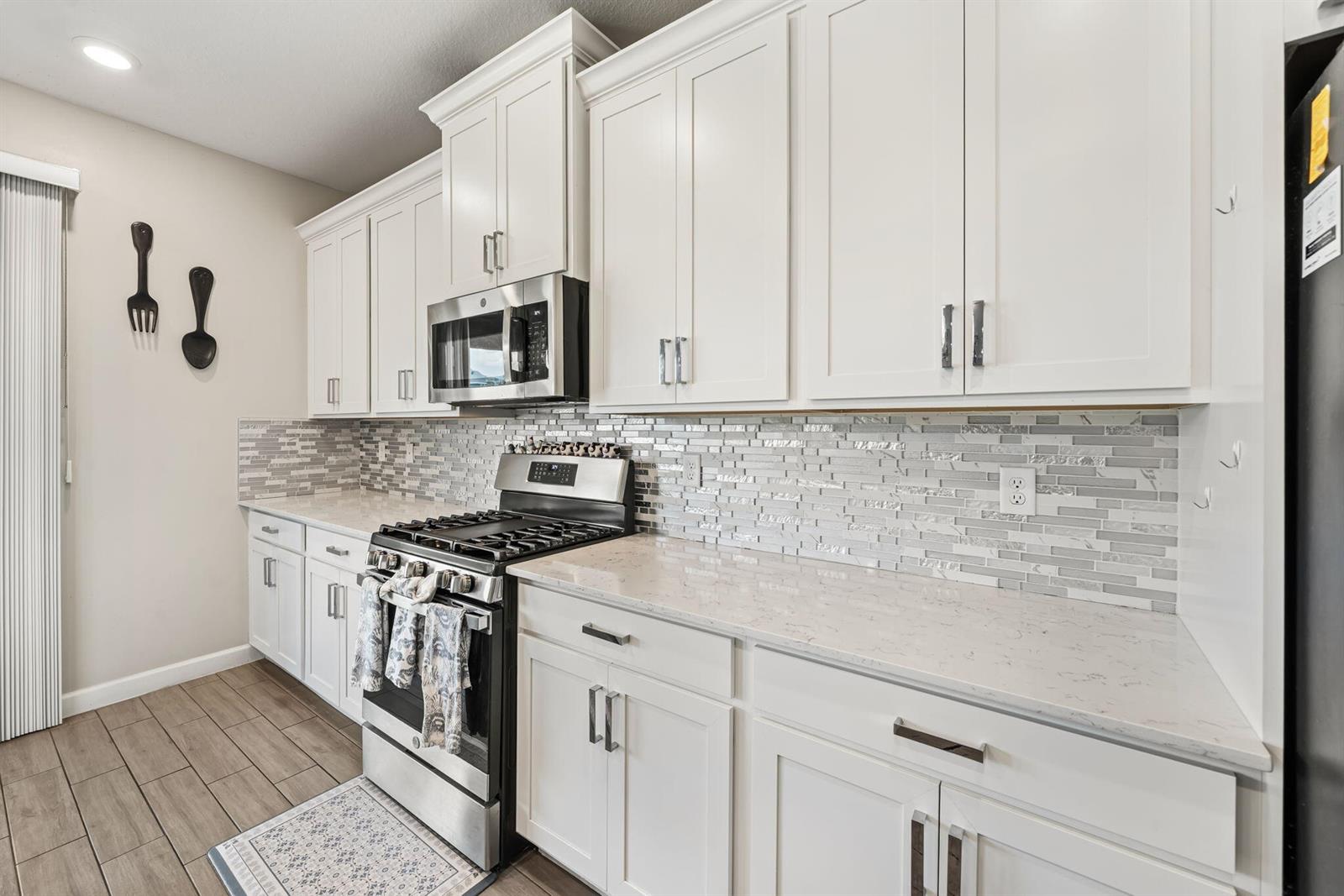 A kitchen features white cabinets, stainless steel appliances, quartz countertops, and a gray tiled backsplash — 1408 Wandering Willow Way, Loxahatchee FL 33470, LOXAHATCHEE, FL
