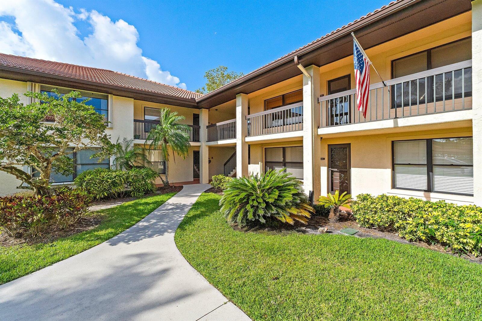 A two-story apartment building exterior with a concrete walkway, landscaping, and an American flag — 671 SW South River Drive # 105, Stuart FL 34997, STUART, FL