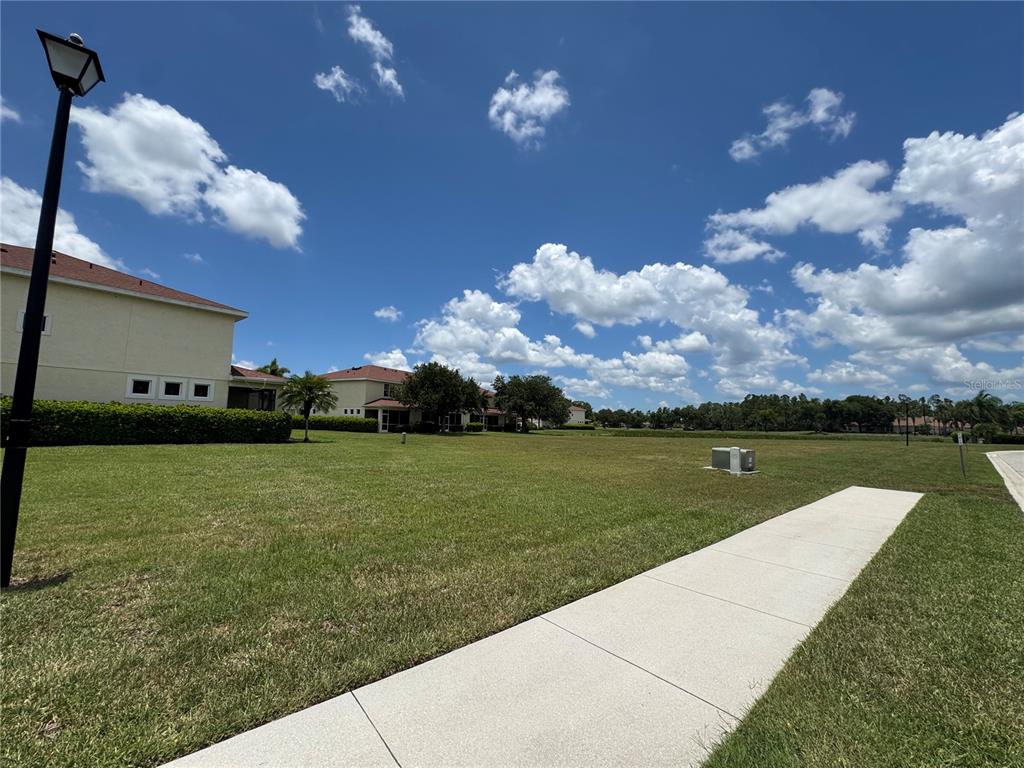 A grassy lawn extends toward residential buildings under a bright blue sky with clouds — JONAH DRIVE, NORTH PORT, FL