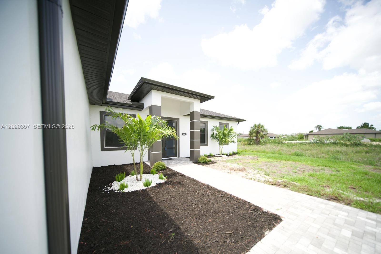 The exterior of a modern white house features dark trim, a paved walkway, and landscaping — 3114 39TH ST SW, Lehigh Acres FL 33976, LEHIGH ACRES, FL