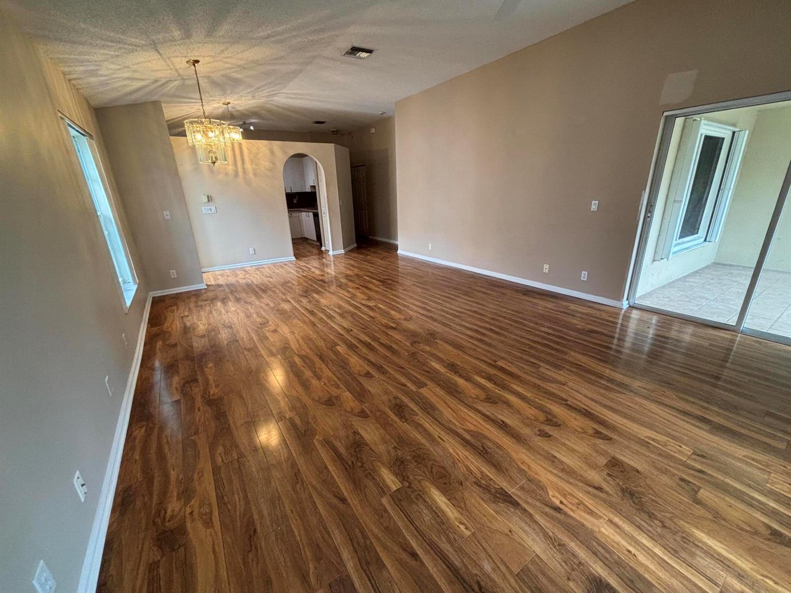 An empty living room features wood-look flooring, beige walls, a crystal chandelier, and sliding glass doors — 8410 Cargill Point, West Palm Beach FL 33411, WEST PALM BEACH, FL