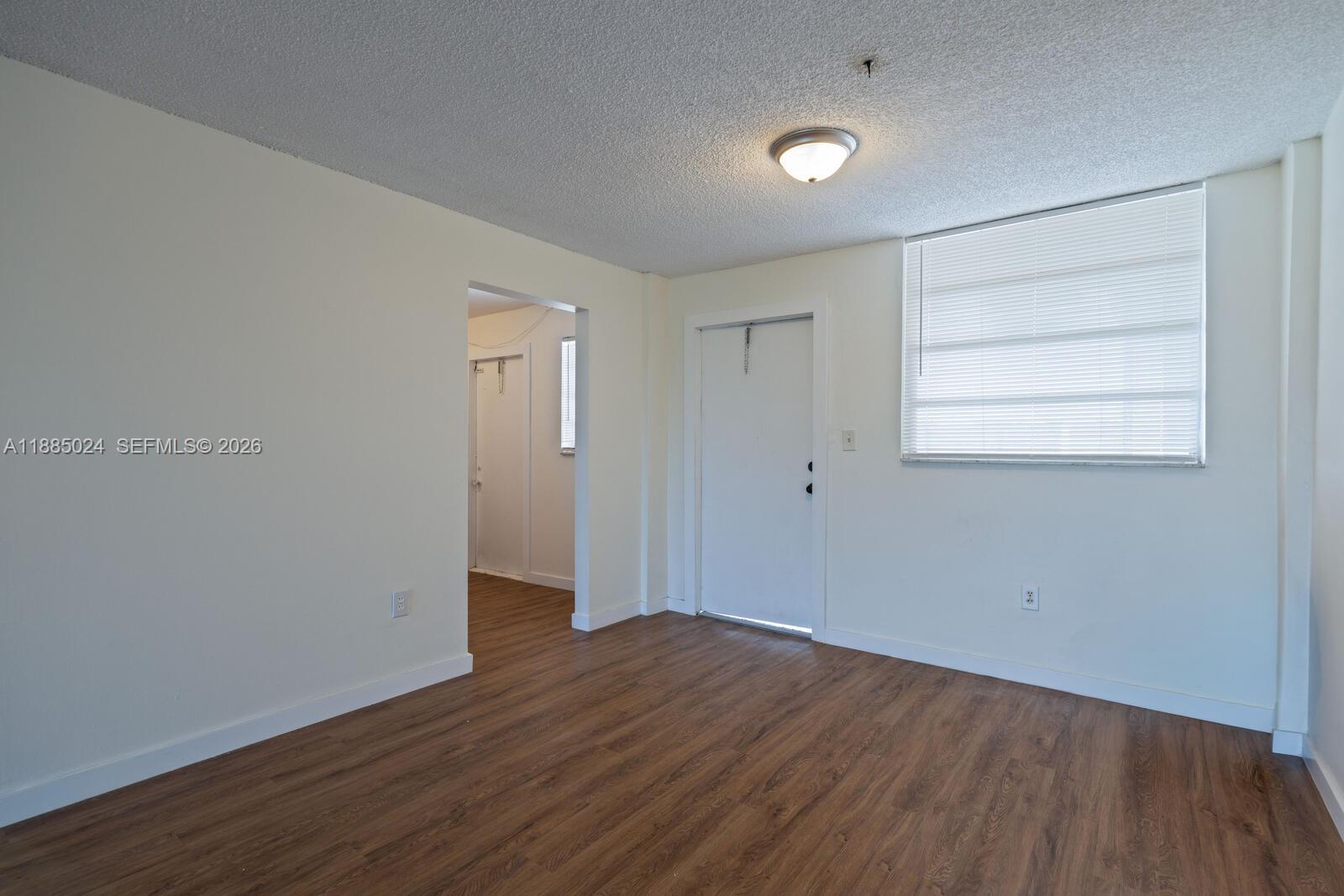 An empty room features wood-look flooring, white walls, a ceiling light, and a window — 14180 SW 84th St # G, Miami FL 33183, MIAMI, FL