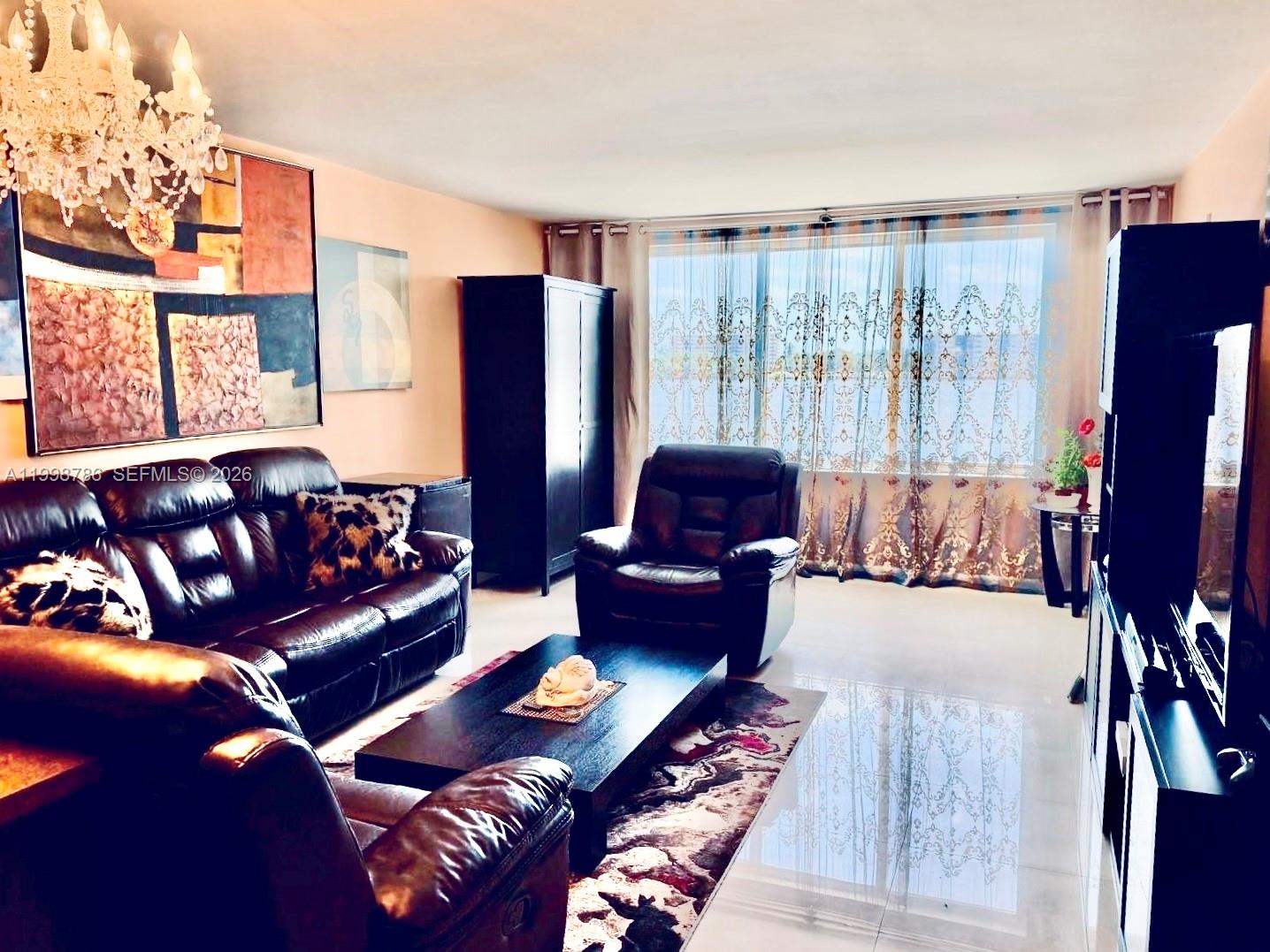 A living room features dark leather furniture, a chandelier, and large windows with patterned curtains — 1150 NE 191st St # 38, Miami FL 33179, MIAMI, FL