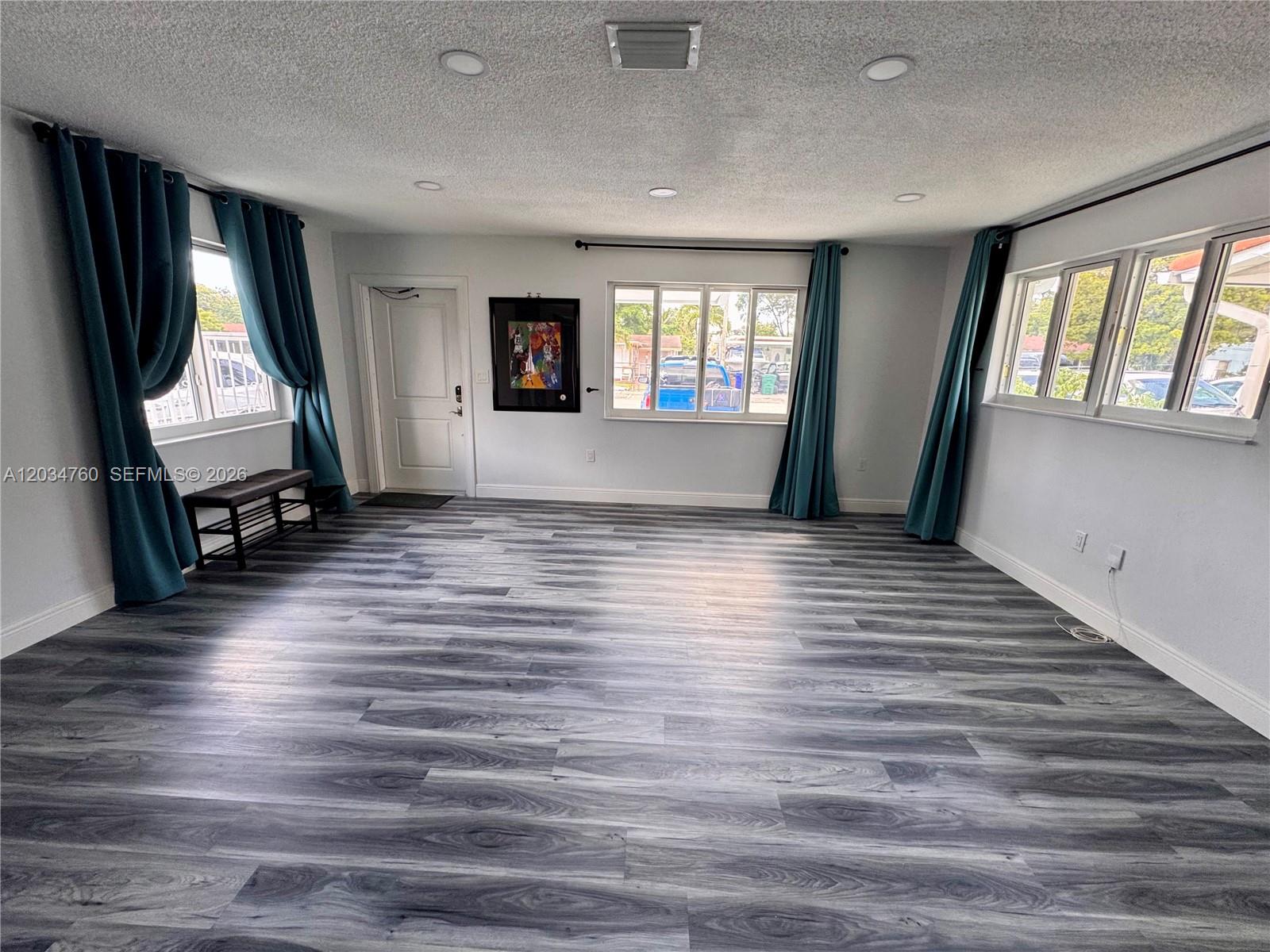 A spacious room features gray wood-look flooring, white walls, recessed lighting, and teal curtains — 1158 NW 34th Ave # 1, Miami FL 33125, MIAMI, FL