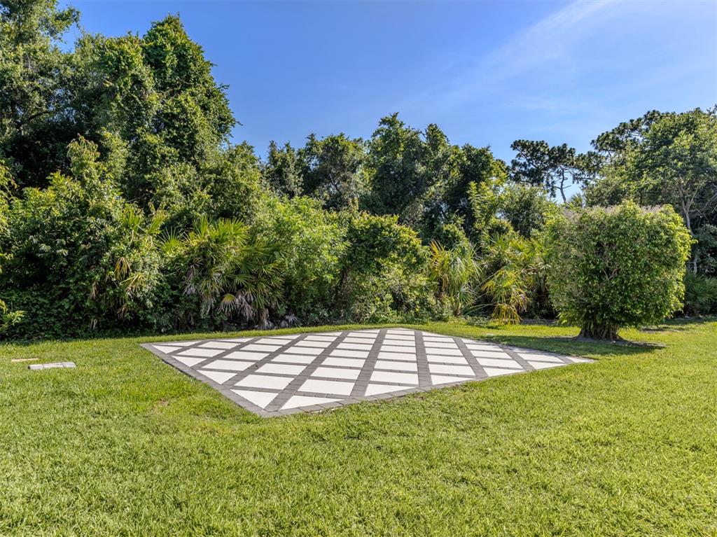 A rectangular stone patio sits in a grassy backyard bordered by dense trees and shrubs — 6511 WESTWARD PLACE, BRADENTON, FL