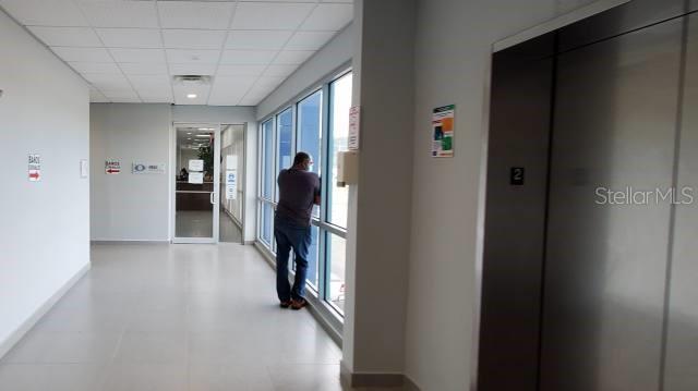 A person stands by a large window in a hallway with a metal elevator door — 1 PR-1 INT. SAKURA ST., CAGUAS, PR