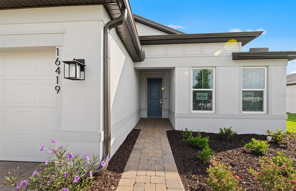 The exterior of a modern house features a paved walkway leading to a blue front door — 16419 LILY ELAINE WAY, BRADENTON, FL