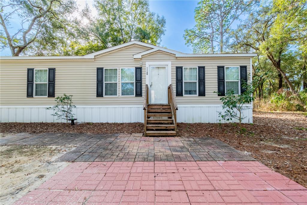 A tan manufactured home features a front entrance with wooden stairs and a paved patio — 11120 NW 107TH TERRACE, CHIEFLAND, FL