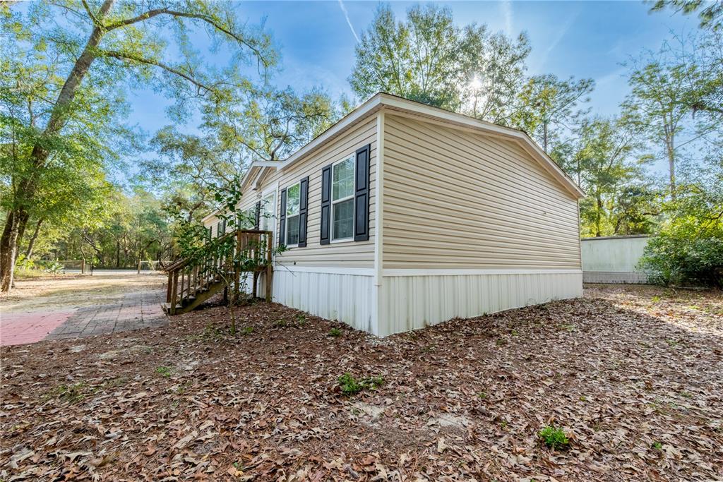 A tan mobile home with white skirting and black shutters sits on a wooded lot — 11120 NW 107TH TERRACE, CHIEFLAND, FL