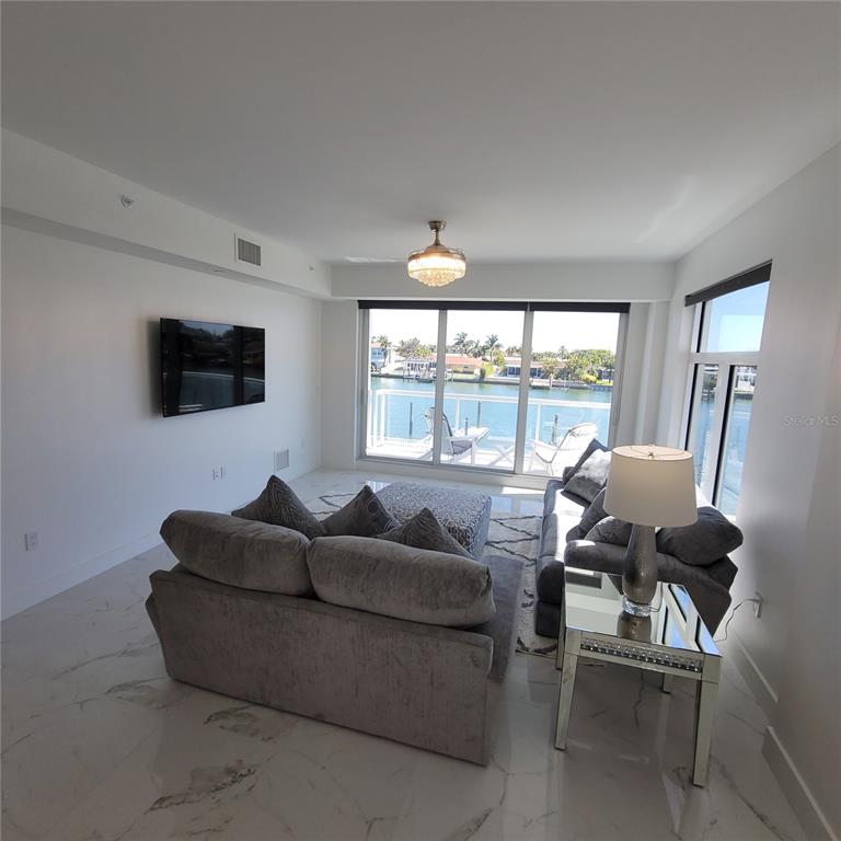 A living room features gray sofas, a mirrored side table, and floor-to-ceiling windows overlooking water — 415 ISLAND WAY UNIT 201, CLEARWATER BEACH, FL