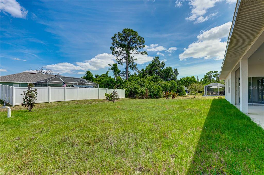 A grassy backyard features a white vinyl fence, a covered patio, and mature trees — 5330 IOTA COURT, NORTH PORT, FL