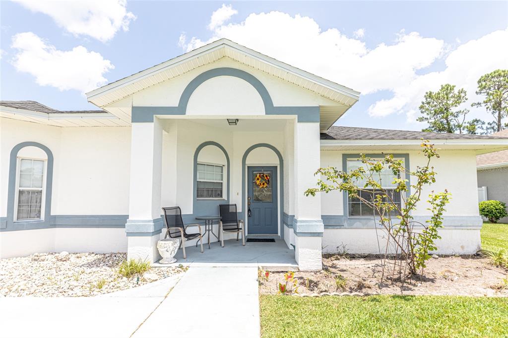 A white house exterior features a covered entryway with a blue door and two chairs — 103 EASTWOOD DRIVE, PALM COAST, FL