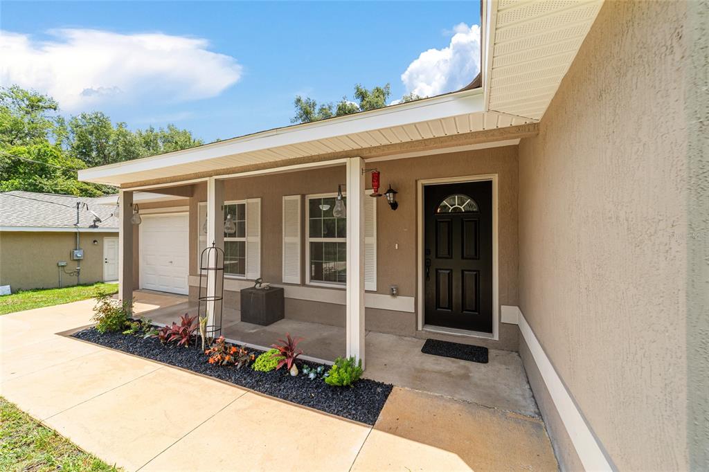 A tan house exterior features a covered front porch, black door, and landscaped garden bed — 2854 SE 140TH LANE, SUMMERFIELD, FL