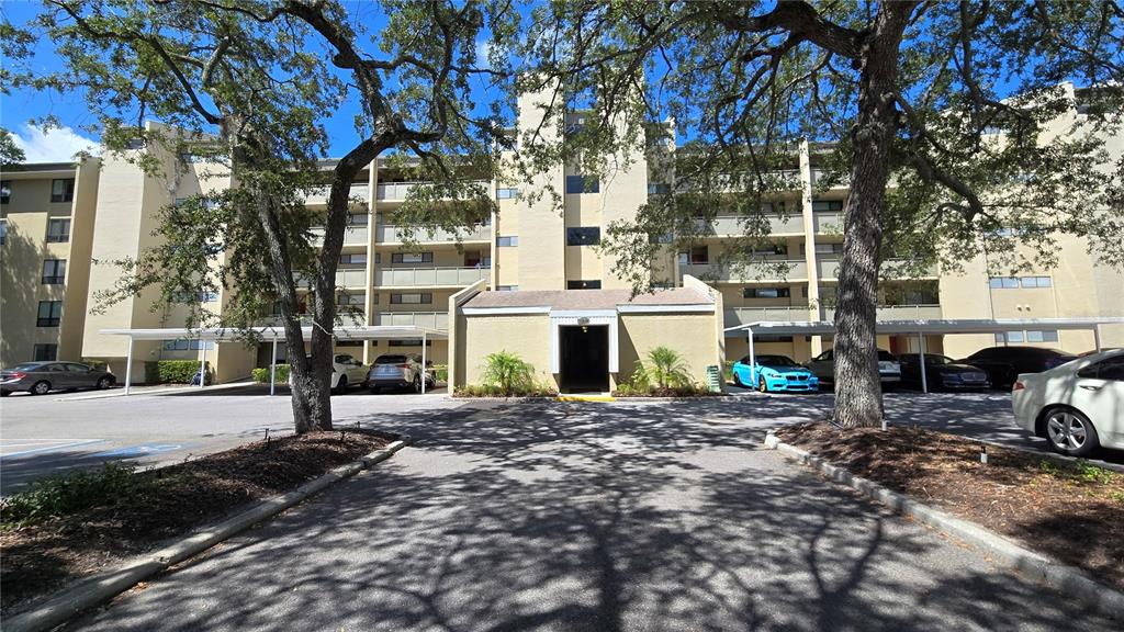 A multi-story apartment building with covered parking and large trees in the foreground — 13626 GREENFIELD DRIVE UNIT 201, TAMPA, FL