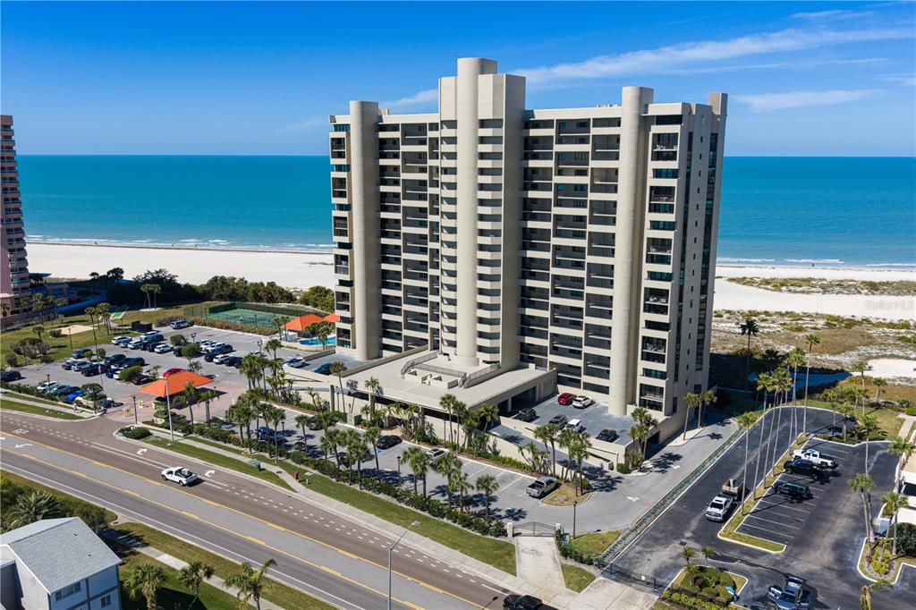An aerial view shows a tall beachfront condominium building with a parking lot and pool — 1290 GULF BOULEVARD UNIT 1002, CLEARWATER, FL