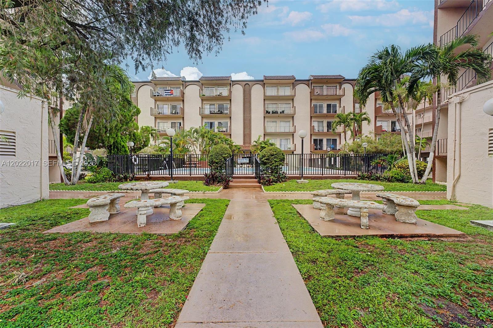 A concrete walkway leads to a fenced pool area with stone tables and apartment buildings — 1950 W 54th St # 105, Hialeah FL 33012, HIALEAH, FL