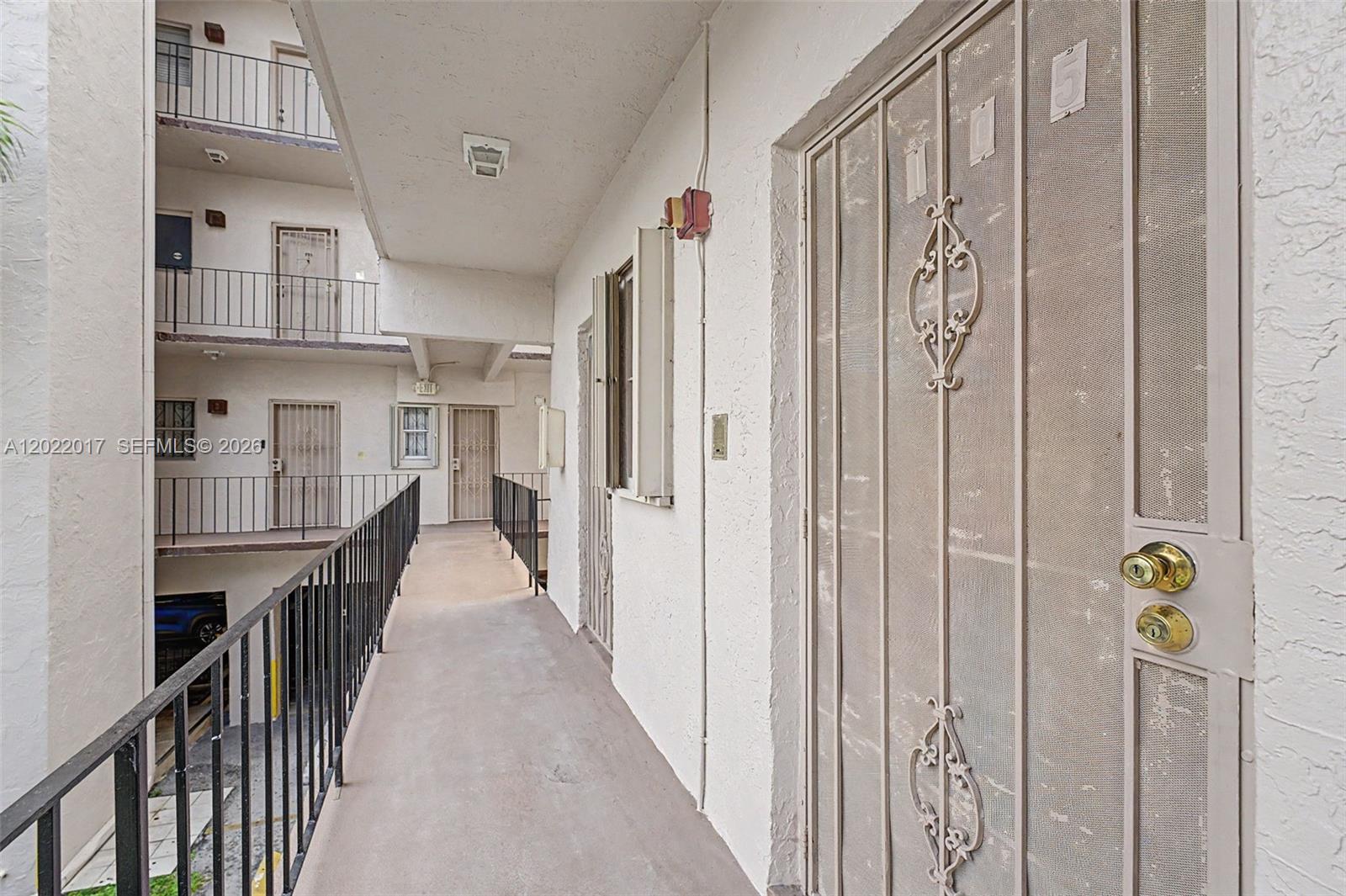 An outdoor apartment building walkway features white stucco walls, metal railings, and security screen doors — 1950 W 54th St # 105, Hialeah FL 33012, HIALEAH, FL