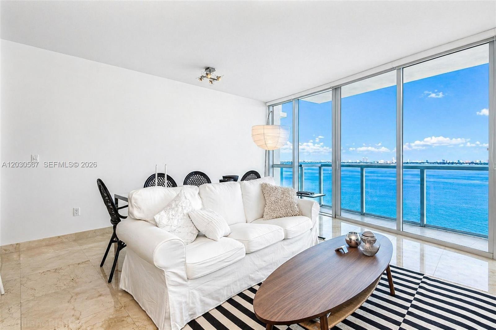 A living room features a white sofa, oval coffee table, and floor-to-ceiling windows overlooking water — 665 NE 25th St # 1202, Miami FL 33137, MIAMI, FL