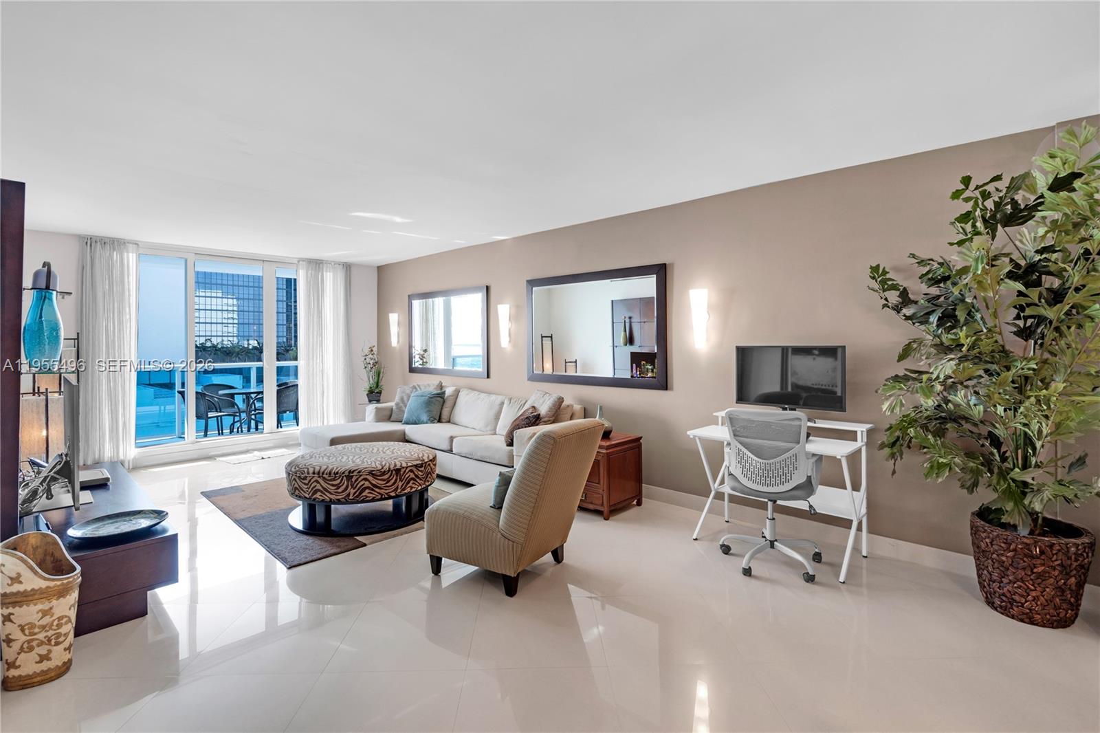A living room features a beige sofa, a patterned ottoman, a desk, and a balcony view — 2301 Collins Ave # 1006, Miami Beach FL 33139, MIAMI BEACH, FL