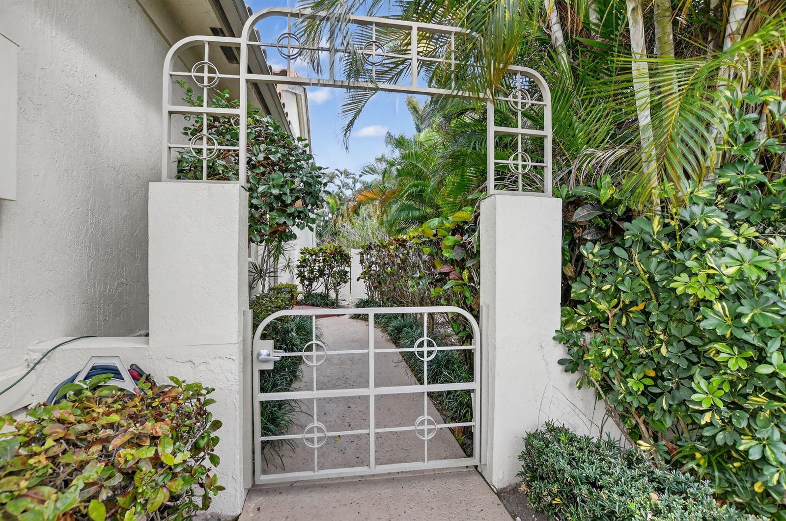 A metal gate and arched trellis frame a concrete walkway leading through a lush garden — 5184 NW 25th Way, Boca Raton FL 33496, BOCA RATON, FL