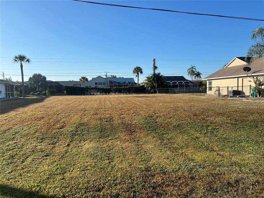 A large, flat, grassy vacant lot sits between residential houses under a clear blue sky — 25285 PADRE LANE, PUNTA GORDA, FL