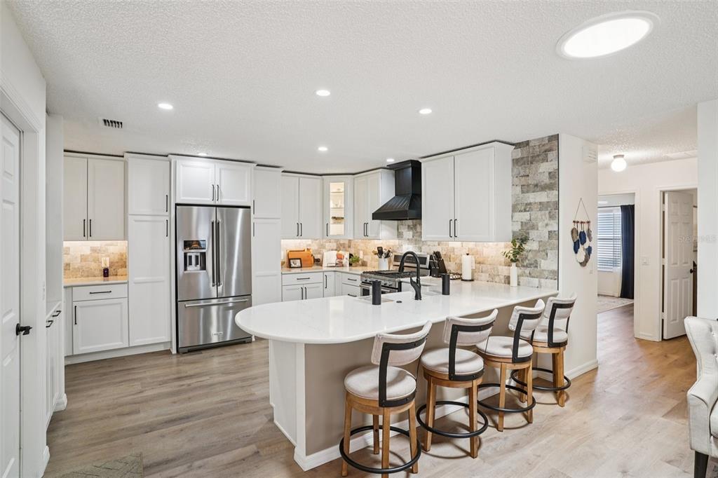 A modern kitchen features white cabinetry, a curved quartz island with bar stools, and stone backsplash — 1609 GIBBES WAY, THE VILLAGES, FL