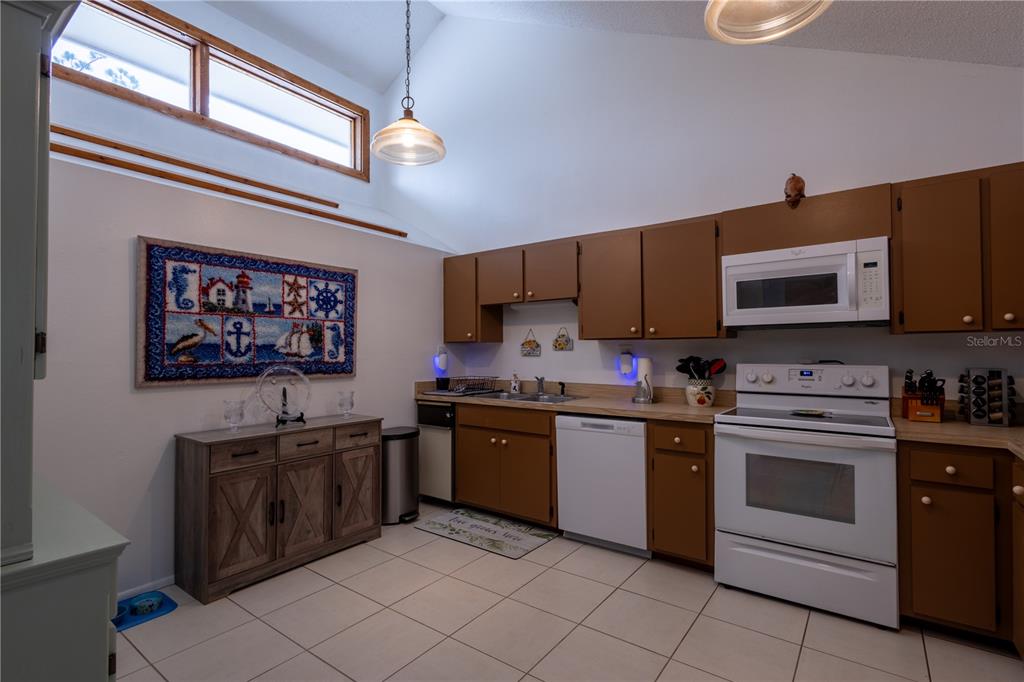 A kitchen with brown cabinets, white appliances, tile flooring, and a vaulted ceiling — 7913 PINEGLEN COURT UNIT 34, SARASOTA, FL