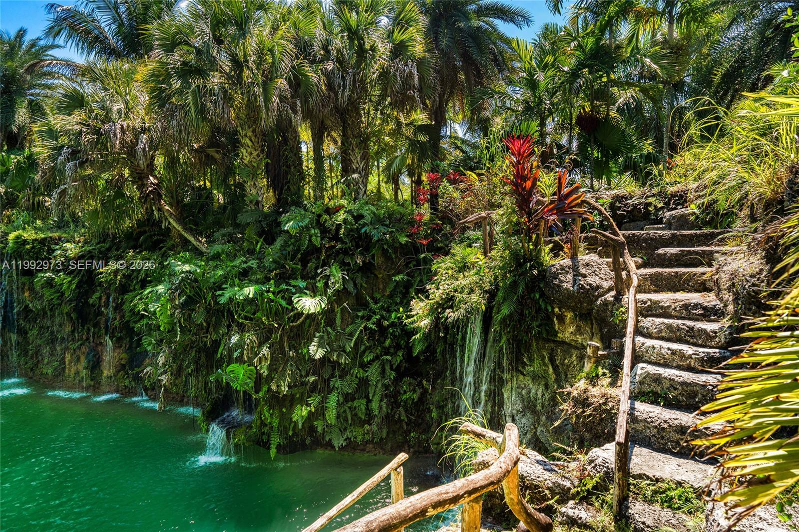 A stone staircase descends alongside a lush, tropical waterfall cascading into a green pool — 15000 SW 200th St, Miami FL 33187, MIAMI, FL