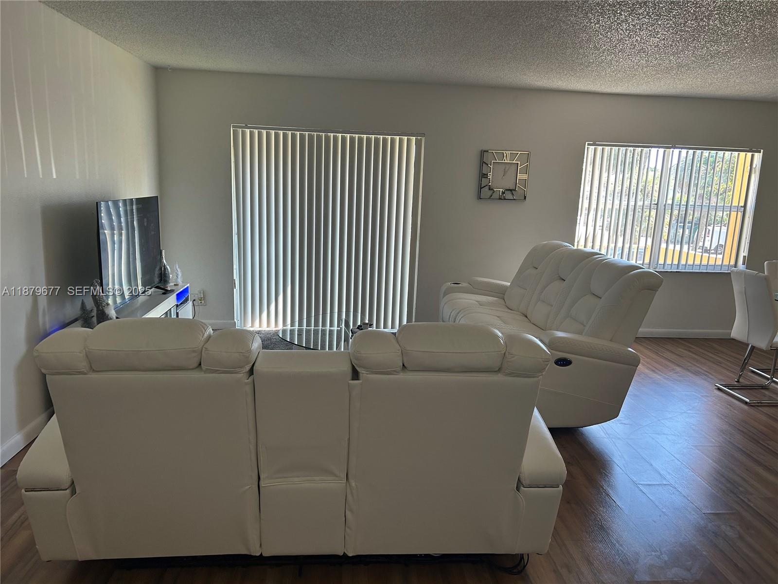 A living room features white leather reclining sofas, wood-look flooring, and vertical window blinds — 17901 NW 68th Ave # R209, Hialeah FL 33015, HIALEAH, FL