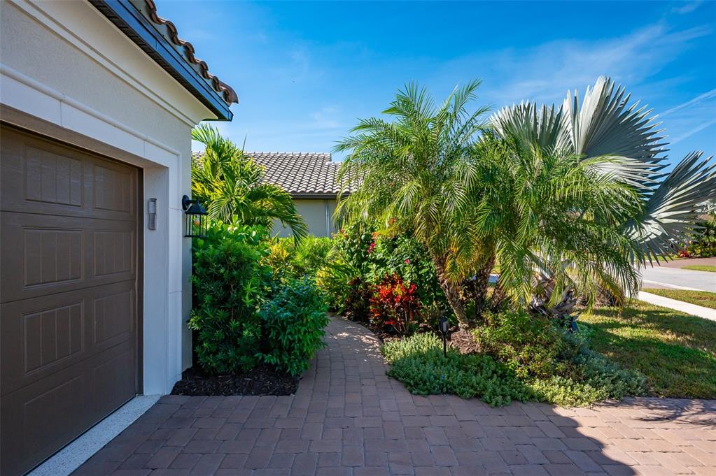A paved walkway leads past a garage and lush tropical landscaping toward a house entrance — 5451 LAGO MAGGIO STREET, SARASOTA, FL