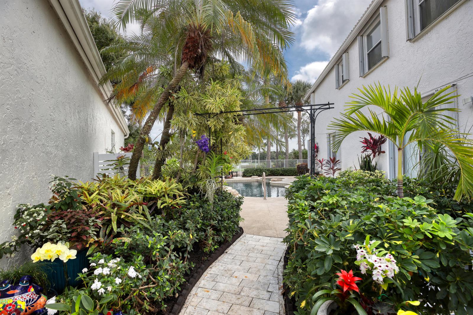 A stone walkway leads through a lush garden toward a swimming pool between two houses — 2070 Chagall Circle, West Palm Beach FL 33409, WEST PALM BEACH, FL
