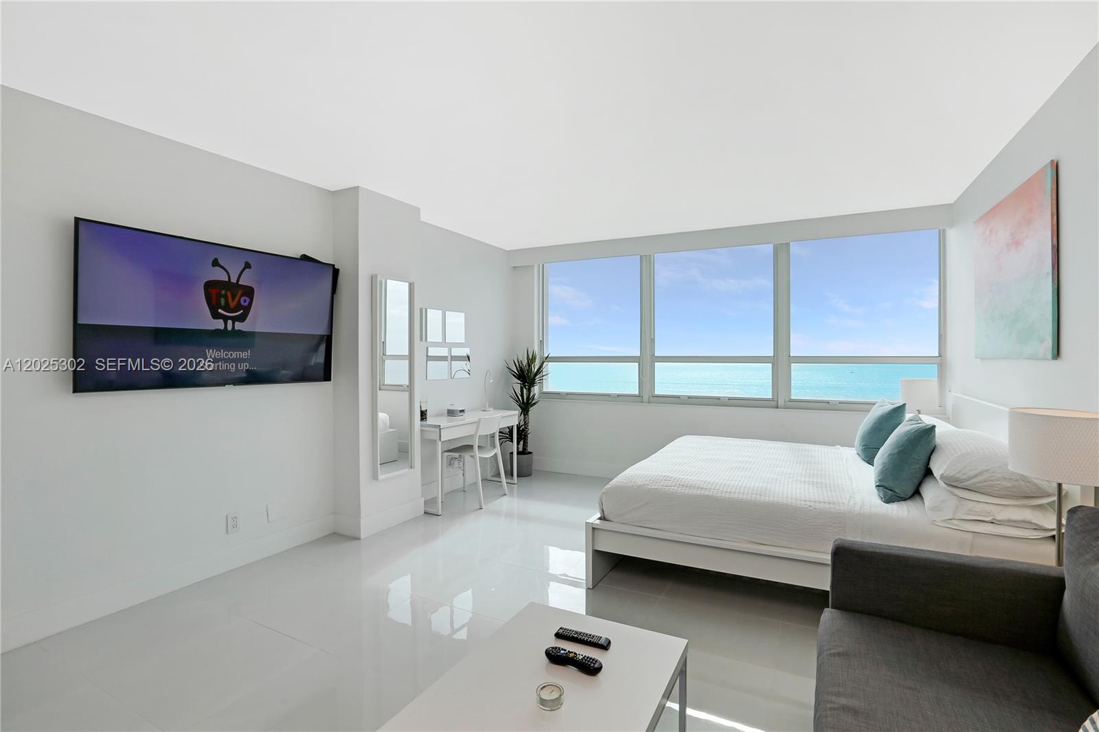 A bright bedroom features a white bed, a wall-mounted television, and floor-to-ceiling ocean views — 5445 Collins Ave # 1203, Miami Beach FL 33140, MIAMI BEACH, FL