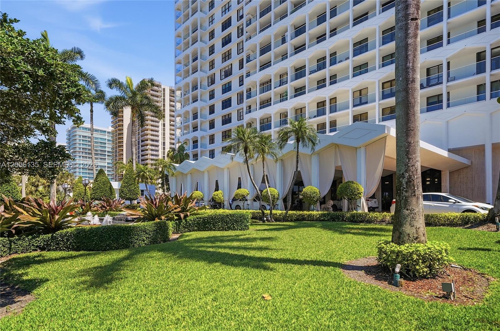 A white high-rise building features a covered entrance with draped fabric and landscaped grounds — 9801 Collins Ave # 14D, Bal Harbour FL 33154, BAL HARBOUR, FL