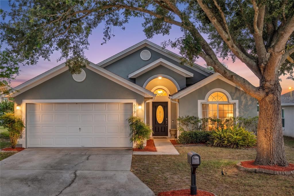 A gray single-story house with a two-car garage, arched entryway, and a large front tree — 659 RESTON PLACE, DAVENPORT, FL